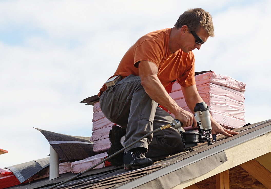 Roofing contractor installing asphalt shingles with a pneumatic nail gun; shingle bundles on the roof.